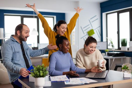 Young diverse group of entrepreneurs with hands up looking surprised at statistic in startup office. Multiethnic business team with laptop and papers excited about project.