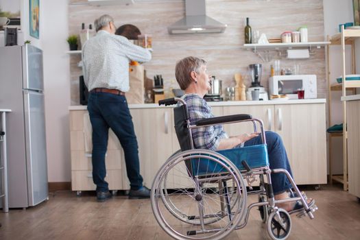 Disabled senior woman sitting in wheelchair in kitchen looking through window. Living with handicapped person. Husband helping wife with disability. Elderly couple with happy marriage.