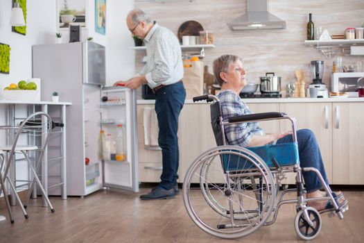 Senior man opening refrigerator while his disabled wife is sitting in wheelchair in kitchen looking through window. Living with handicapped person. Husband helping wife with disability. Elderly couple with happy marriage.