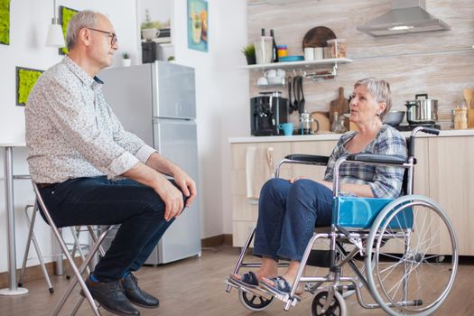 Retired invalid woman in wheelchair having a conversation with old elderly husband in kitchen. Old man talking with wife. Living with disabled person with walking disabilities
