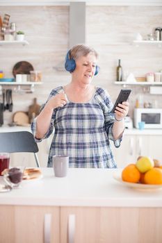 Relaxed senior woman listening music in headphones during breakfast in kitchen. Elderly person dancing, fun lifestyle with modern technology