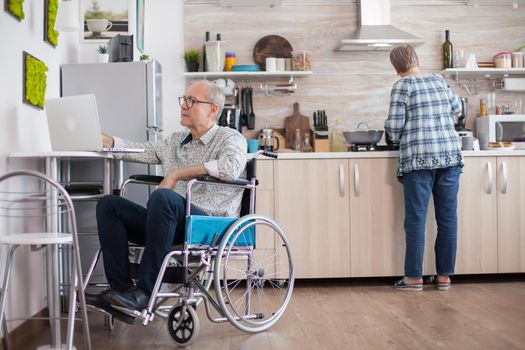 Disabled senior man in wheelchair working on laptop in kitchen while wife is preparing delicious breakfast for both of them. Man using modern technology while working from home.