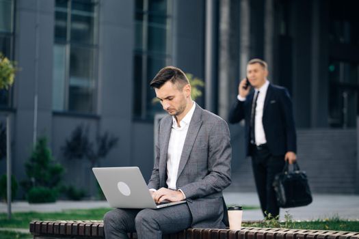 Man in suit working with laptop while sitting on bench. Remote work concept. Distance working. Isolated man on a suit. Businessman typing on laptop computer outdoor.