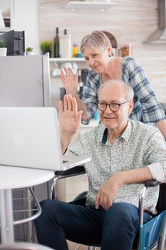 Disabled senior man in wheelchair and his wife having a video conference on laptop in kitchen. Paralyzed old man and his wife having a online conference.