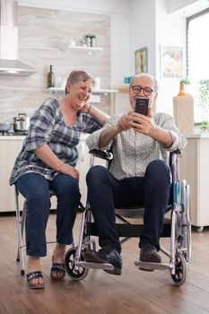 Disabled senior man in wheelchair and his wife laughing and browsing with a modern smartphone in kitchen. Paralyzed old man and his wife having a online conference.
