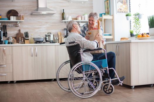 Senior woman taking grocery paper bag from handicapped husband in wheelchair. Mature people with fresh vegetables from market. Living with disabled person with walking disabilities