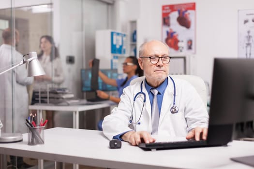 Elderly aged physician with stethoscope working on computer in clinic office while young medic is discussing illness rapport with patient on hospital corridor and nurse is holding sick man x-ray.