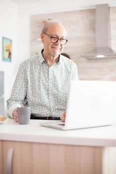 Old man smiling while watching a movie on the laptop in the kitcen. Daily life of senior man in kitchen during breakfast using laptop holding a cup of coffee. Elderly retired person working from home, telecommuting using remote internet job online communication on modern technology notebook