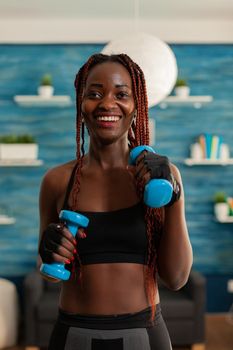 Fit black woman exercising biceps with dumbbells in home living room, dressed in sportwear. Sportive strong active african smiling at camera during workout with healthy lifestyle.