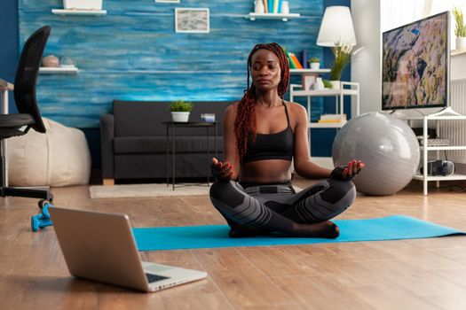 Afro american woman in sportwear sitting practicing lotus position doing yoga sitting on yoga mat dressed in sportwear following online training. Workout and healthy lifestyle.