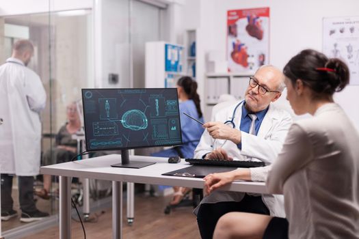 Senior doctor pointing at ct scan on computer in hospital office for young patient. Young medic discussing with disabled old woman in wheelchair on clinic corridor. Nurse wearing blue uniform.