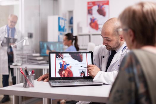 Cardiologist in white coat pointing at heart on laptop screen in hospital office and talking with old woman about treatment for her illness. Mature doctor writing prescription on clipboard in clinic corridor.