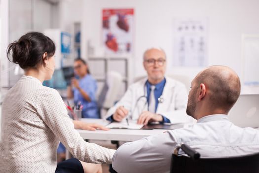 Handicapped man in wheelchair after car accident with wife at hospital for periodic check up with doctor. Senior medic explaining illness to disabled patient. Nurse holding x-ray image.