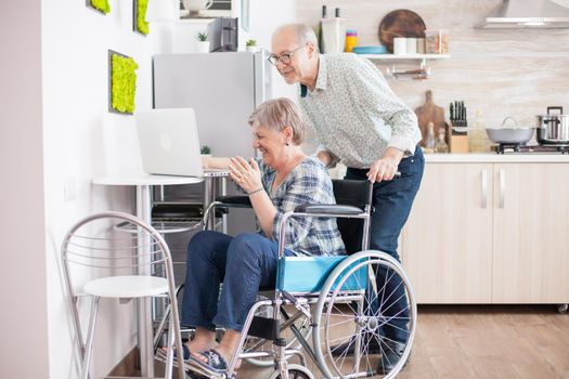 Senior couple laughing during a video call with grandchildrens using tablet computer in kitchen. Paralyzed handicapped old elderly woman using modern communication techonolgy.