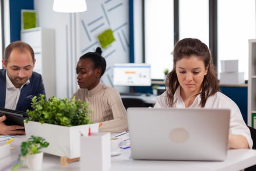 Focused woman manager typing on laptop, browsing on internet while sitting at desk concentrated having multitasks. Diverse colleagues working in background. Multiethnic coworkers planning new financial project.