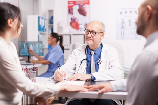 Senior doctor writing notes on clipboard during examination of young couple with fertility illness in hospital office and nurse holding patient x-ray.