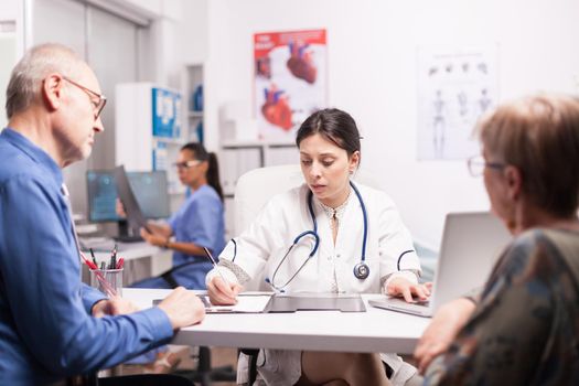 Young doctor writing prescription for sick senior couple during medical examination in hospital office. Medic wearing medical stethoscope and nurse blue uniform.