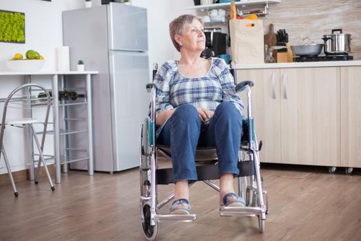 Handicapped senior woman in wheelchair standing alone in kitchen looking throug window. Pensive thoughtful lonely woman in solitude. Elderly disabled pensioner after injury and rehab, paralysis and disability depressed invalid full of sorrow, worry and sad face. Retirement period for old people