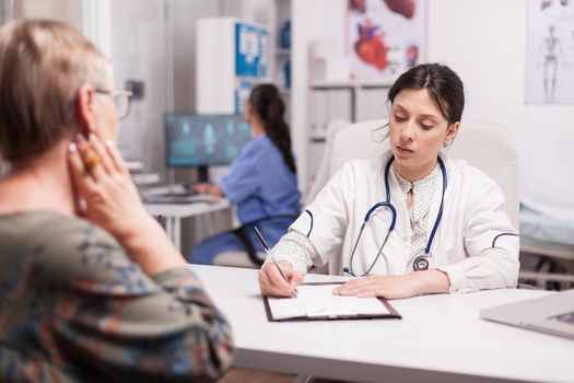 Doctor in hospital office writing treatment for senior woman during examination. Nurse wearing blue uniform working on computer.