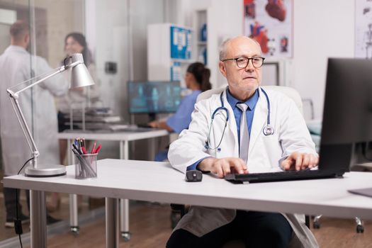 Senior man doctor with grey hair working on computer while young colleague is with a patient on hospital corridor and nurse checking analysis report.