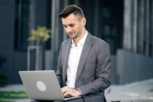 Businessman typing on laptop computer outdoor. Man in suit working with laptop while sitting on bench. Remote work concept. Young businessman in the city sits on bench, takes a laptop and works.