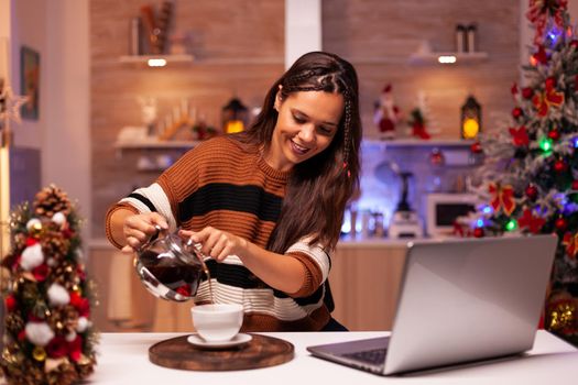 Modern woman filling mug of tea from kettle on video call connection talking to friends. Cheerful young person using laptop for wishing merry christmas in seasonal decorated kitchen