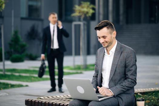Distance working. Isolated man on a suit. Caucasian businessman in suit and tie communication while typing on laptop sitting outside