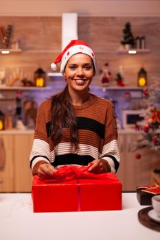 Cheerful woman decorating gift boxes using ribbon and wrapping paper for christmas eve party night. Festive young person with santa hat in holiday decorated kitchen waiting for celebration