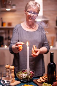 Senior woman with glasses lighting candle on kitchen table for romantic dinner. Elderly woman waiting her husband for a romantic dinner. Mature wife preparing festive meal for anniversary celebration.