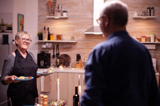 Elderly wife smiling at senior husband in kitchen during romantic dinner. Elderly old couple talking, sitting at the table in kitchen, enjoying the meal, celebrating their anniversary.