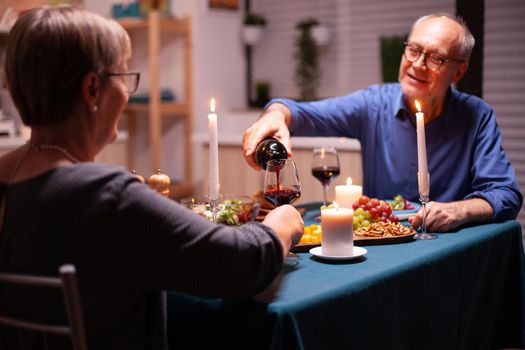 Senior man chatting with wife and pouring red wine. Elderly couple sitting at the table in kitchen, talking, enjoying the meal, celebrating their anniversary in the dining room.