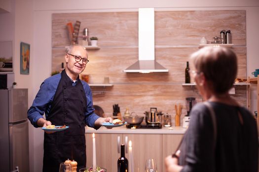 Old man talking with wife while serving her dinner during relationship celebration. Senior couple talking, sitting at the table in kitchen, enjoying the meal, celebrating their anniversary in the dining room.