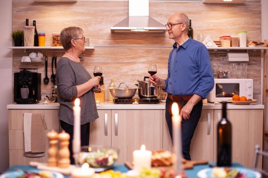 Senior old couple drinking wine an talking during romantic dinner in kitchen. Aged couple in love talking having pleasant conversation during healthy meal.