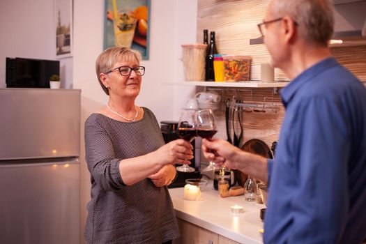 Mature couple looking at each other holding glasses of red wine in the evening. Aged couple in love talking having pleasant conversation during healthy meal.