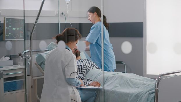 Practitioner specialist doctor writing sickness treatment on clipboard monitoring disease symptom discussing medical expertise. Woman nurse arrange patient bed in hospital ward during rehabilitation