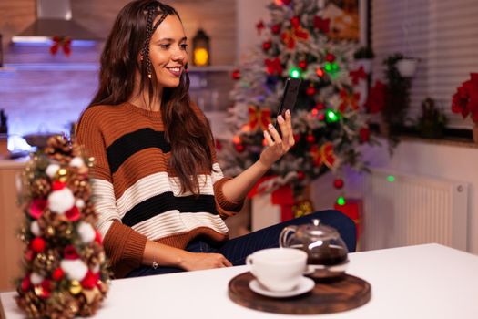 Adult woman with smartphone for video call conference on online internet and webcam with friends in kitchen decorated with ornaments and christmas lights for seasonal winter celebration