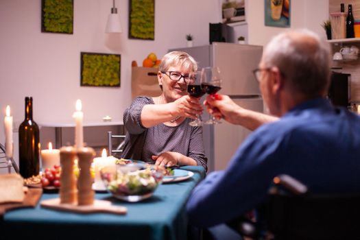 Handicapped man in wheelchair toasting with wife during dinner. Happy cheerful senior elderly couple dining together in the cozy kitchen, enjoying the meal, celebrating their anniversary.