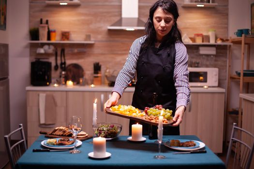 Woman holding wooden plate with mix of grapes and cheese while cooking romantic dinner. Young caucasian lady cooking for her husband a romantic dinner, waiting in kitchen.