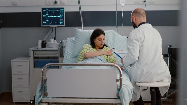 Caucasian female with nasal oxygen tube sitting in bed in hospital ward. Practitioner doctor discussing with patient about medical expertise writing disease test results on clipboard