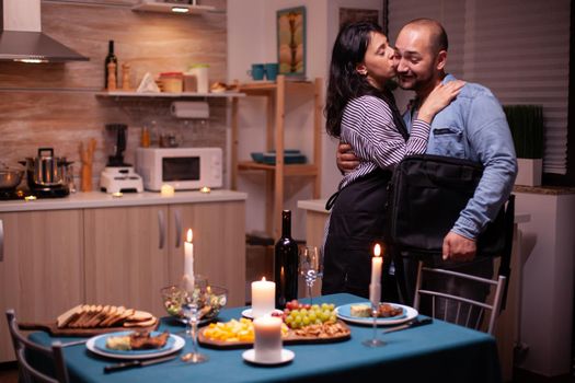 Kissing husband cheek while greeting him in kitchen for romantic dinner. Woman preparing festive dinner with healty food, cooking for his man romantic dinner, talking, sitting at table