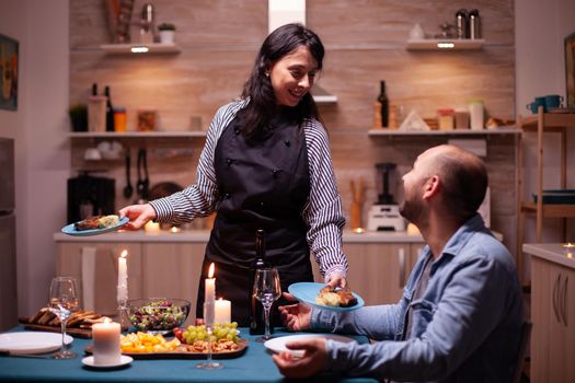 Delighted husband and wife having romantic dinner with tasty food in kitchen. Woman preparing festive dinner with healthy food, cooking for his man romantic dinner, talking, sitting at table