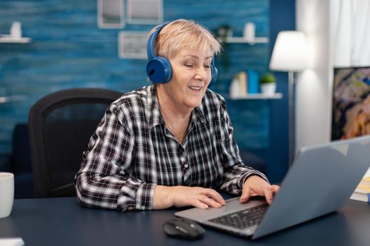 Mature lady listening music smiling and entertaining herself Retired senior woman wearing headphones in the course of using laptop computer in home living room.