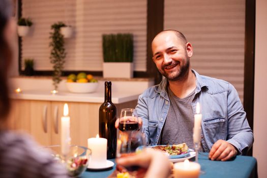Boyfriend smiling at wife enjoying anniversary celebration in kitchen. Talking happy sitting at table dining room, enjoying the meal at home having romantic time at candle lights.