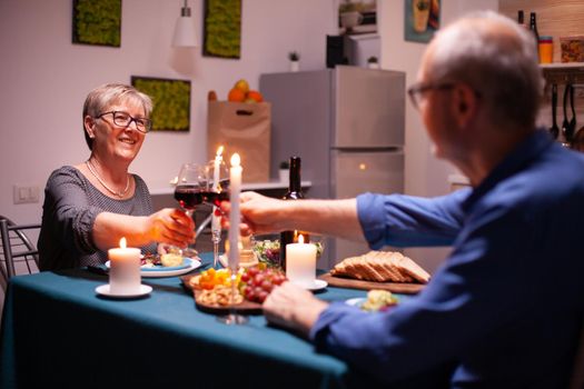 Happy pensioner couple toasting with red wine glasses in kitchen. Senior couple sitting at the table in kitchen, talking, enjoying the meal, celebrating their anniversary in the dining room.