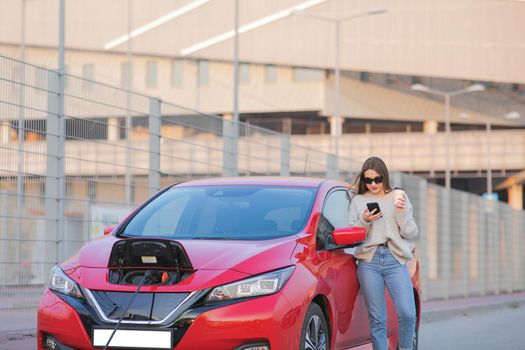 Stylish girl stands with phone near her red electric car and waits when vehicle will charged. Connecting the Charger Plug of an Electric Car