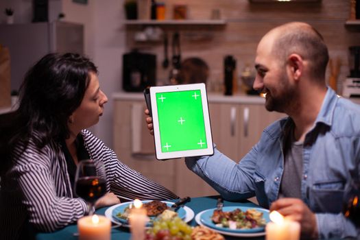 Couple using tablet pc with chroma key and enjoying their time together during romantic dinner. Husband and wife looking at green screen template chroma key display sitting at the table in kitchen during dinner.