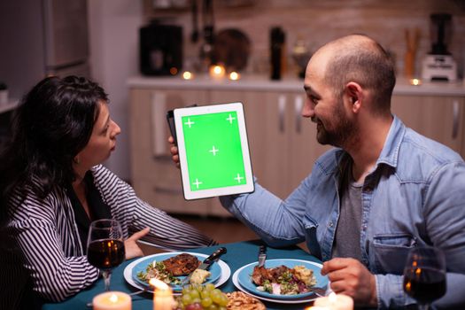 Couple using tablet pc with green template during festive dinner. Husband and wife looking at green screen template chroma key display sitting at the table in kitchen.