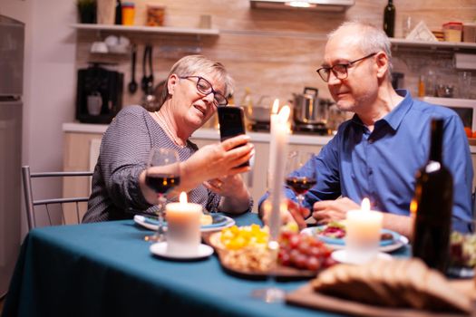 Elderly woman using smartphone to show her husband photos during romantic dinner in kitchen. Sitting at the table in the kitchen, browsing, searching, using phone, internet, celebrating their anniversary in the dining room.