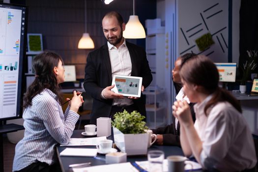 Entrepreneur businessman showing company strategy using tablet for corporate presentation working overtime in meeting office room late at night. Diverse multi-ethnic teamwork brainstorming ideas.