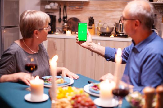 Old retired senior couple holding mockup phone at dinner. Aged people looking at mockup template chroma key isolated smart phone display using techology internet sitting at the table in kitchen.